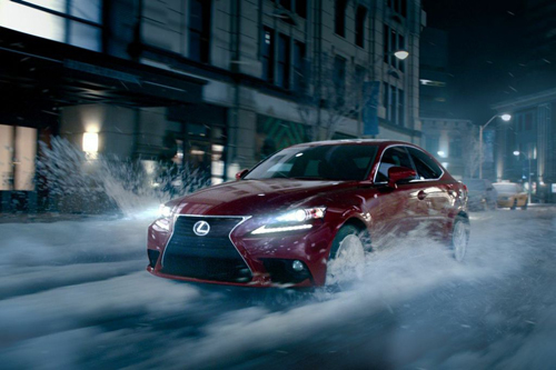 A red Lexus sedan driving through deep snow on a city street at night, splashing snow into the air.