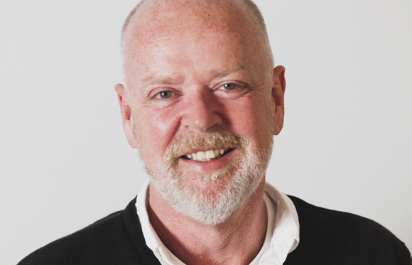 A smiling middle-aged man with a grey beard and blue eyes wearing a white shirt and black sweater.