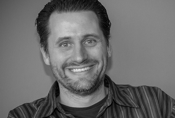 Black and white headshot of a smiling man with short dark hair and a beard wearing a striped shirt.