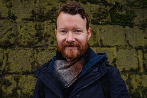 A man with reddish hair and a beard wearing a scarf and dark jacket in front of a stone wall.