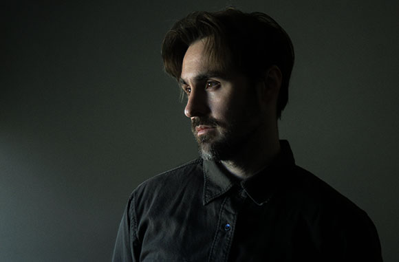 Side profile of a man with dark hair and a beard, lit from the side against a dark background.