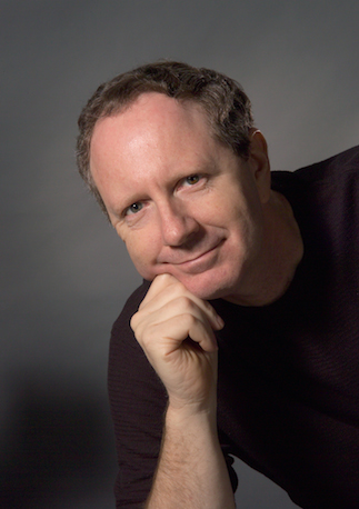 A man with short brown hair and a black shirt rests his chin on his closed fist against a gray background.