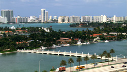 Aerial view of a city skyline with high-rise buildings, a bridge crossing a blue waterway, and lush green islands.