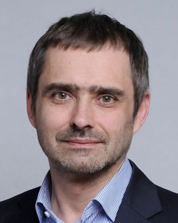 A man with short brown hair and light stubble wearing a blue striped shirt and dark blazer.