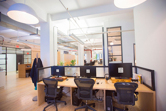 Modern office interior with wooden desks, computer monitors, black office chairs, and bright overhead lighting.