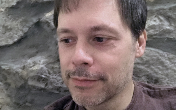 A man with short dark hair and light stubble looking to the left against a textured stone background.
