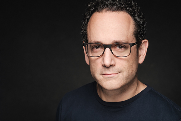 A man with dark curly hair and glasses looking forward against a dark background.