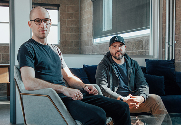 Two men sit in chairs in a room with large windows and brick walls.