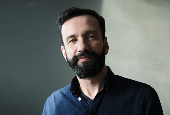 A man with dark hair and a full black beard looking toward the camera against a neutral background.