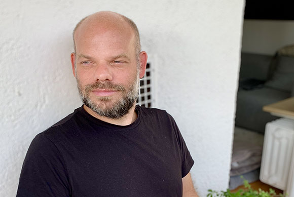 A bald man with a salt-and-pepper beard wearing a black t-shirt, looking off to the side.