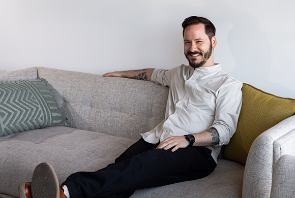 A bearded man in a white button-down shirt sits on a gray couch with his arm resting on the backrest.