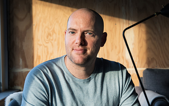 A bald man wearing a light grey sweater sits in front of a wooden wall in warm sunlight.
