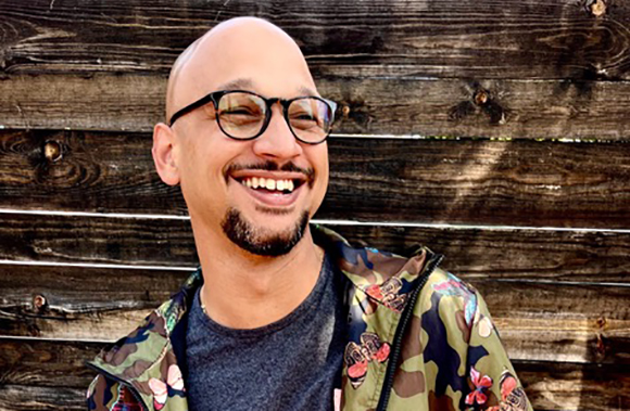 A bald man with glasses and a goatee smiles while wearing a camouflage jacket against a wooden wall.