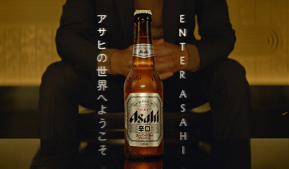 A brown glass bottle of Asahi Dry beer stands in front of a person sitting in a dark room.