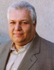 A middle-aged man with short gray hair wearing a tan collared shirt and a gray blazer.