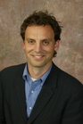 A smiling man with dark wavy hair wearing a blue shirt and a black blazer.