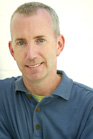 A man with short light hair and blue eyes smiling while wearing a blue polo shirt.