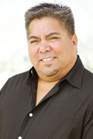 A smiling man with short dark hair and a goatee wearing a black collared shirt.