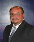 A man with glasses, a mustache, and a goatee wearing a dark suit, white shirt, and light blue tie.