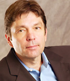 A man with brown hair wearing a dark blazer and a light blue button-down shirt.
