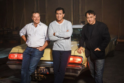 Three men standing in front of a light yellow vintage car in a dimly lit garage.