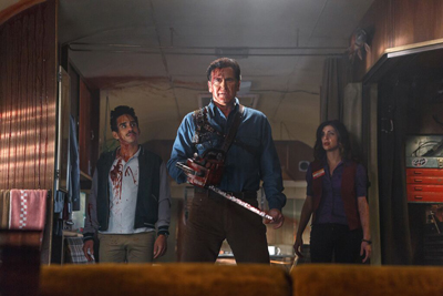 A man holding a chainsaw stands between a man and a woman in a dark trailer.