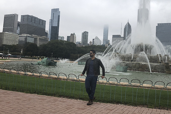 A man in a leather jacket stands before a large, spraying fountain with skyscrapers in the background.