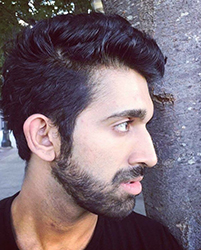 Side profile of a man with dark wavy hair and a beard leaning against a tree.