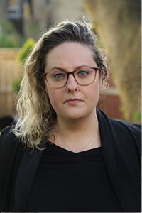 A woman with wavy blonde hair and red-rimmed glasses looking forward against a blurred outdoor background.