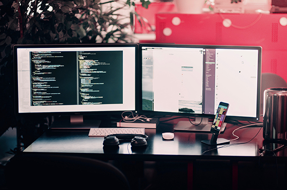 Two computer monitors on a desk showing programming code and application windows in a dimly lit room.
