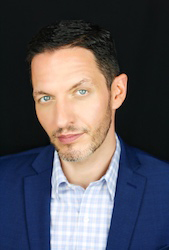 A man with short dark hair and light eyes wearing a blue blazer and a light blue checkered shirt.