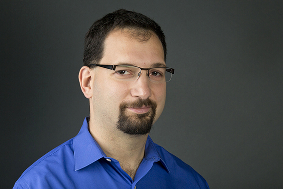 A man with dark hair, glasses, and a goatee wearing a blue button-down shirt against a dark background.