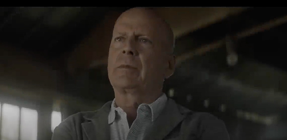 A bald man in a grey jacket looking upward with a solemn expression in a dimly lit setting.