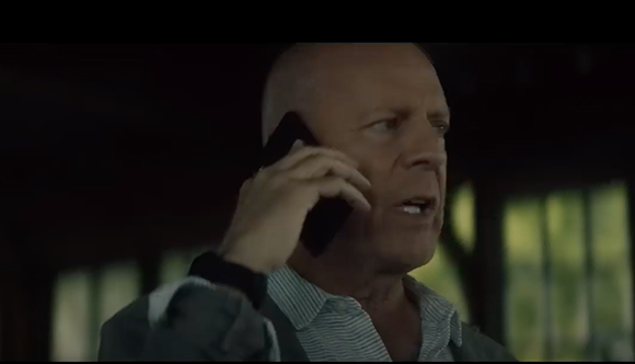 A bald man in a striped shirt speaks intensely into a black smartphone.