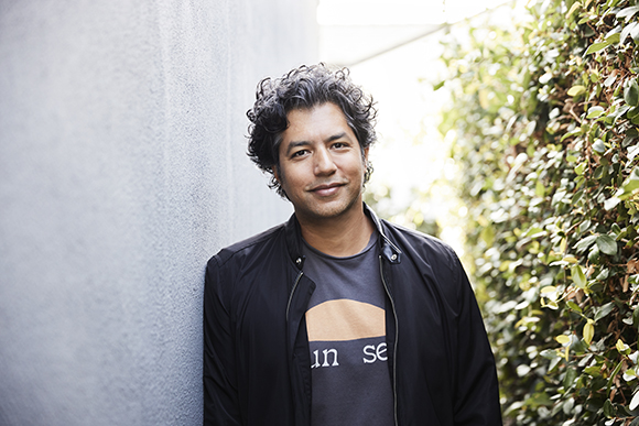 A man with curly dark hair and a black jacket leans against a light gray wall next to green foliage.