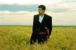 A man in a dark suit and white shirt stands in a golden field under a cloudy sky.