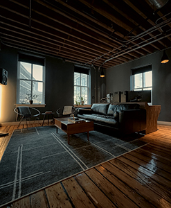 A dark industrial living room with a black leather sofa, wooden coffee table, and large windows.