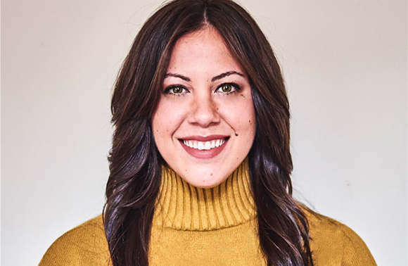 A smiling woman with long dark hair wearing a mustard yellow turtleneck sweater.