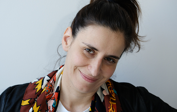 A woman with dark hair in a ponytail smiles and tilts her head against a light background.