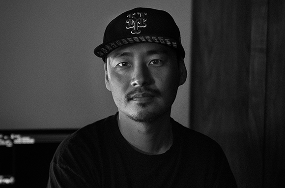 Black and white portrait of a man with a mustache wearing a dark baseball cap with a logo.