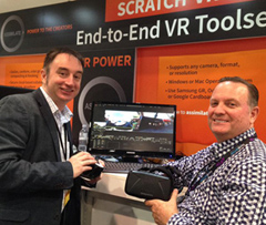 Two men smiling next to a computer monitor and a VR headset at a trade show booth.