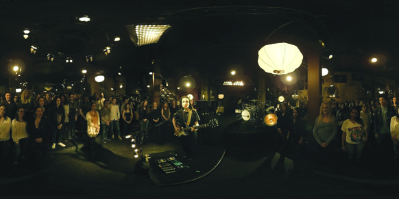 A musician plays an electric guitar on a stage in a dark, crowded club with hanging lanterns.
