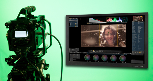 Professional video camera on a tripod next to a monitor showing color grading software with a woman's face.