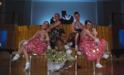 Five women in sparkling, colorful dresses pose together on a stage with gold curtains behind them.
