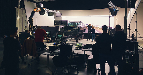 Film crew in a dark studio setting up lighting and cameras around a car on a platform.