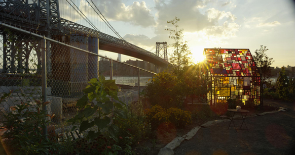 A colorful stained-glass structure in a garden with a large suspension bridge in the background at sunset.