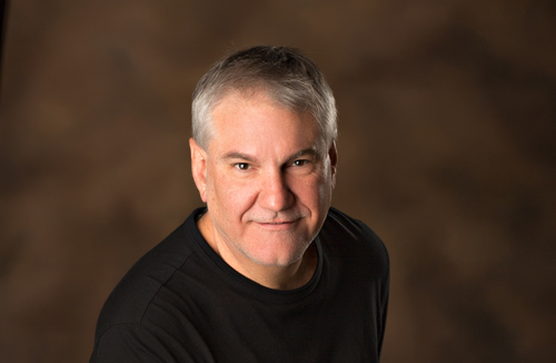 A man with short gray hair and a black t-shirt smiles against a dark brown background.
