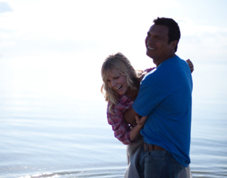 A smiling man in a blue shirt hugs a blonde woman in a pink shirt by the ocean.