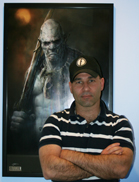 A man in a striped polo shirt stands with arms crossed in front of a large framed poster of a fantasy creature.