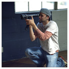 A man in a grey cap and white t-shirt kneels while looking through a camera lens.